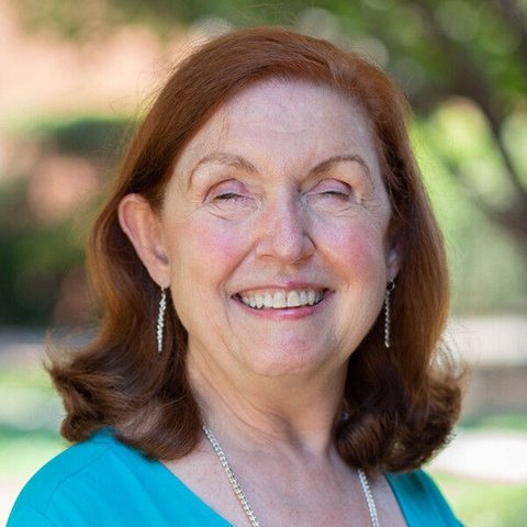 Portrait of Daryle Hines, smiling, with shoulder-length auburn hair, wearing a teal top and silver earrings, outdoors